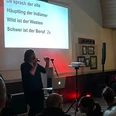 IMG_4478.jpeg Frau mit Mikrofon vor Leinwand mit Text 'Da sprach der alte Häuptling der Indianer Wild ist der Westen schwer ist der Beruf 2x' vor Publikum in dunklem Raum