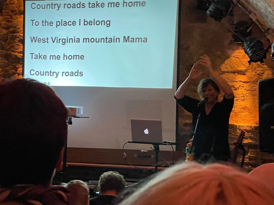 IMG-20240410-WA0008.jpg Frau mit Mikrofon vor Leinwand mit Text 'Da sprach der alte Häuptling der Indianer Wild ist der Westen schwer ist der Beruf 2x' vor Publikum in dunklem Raum