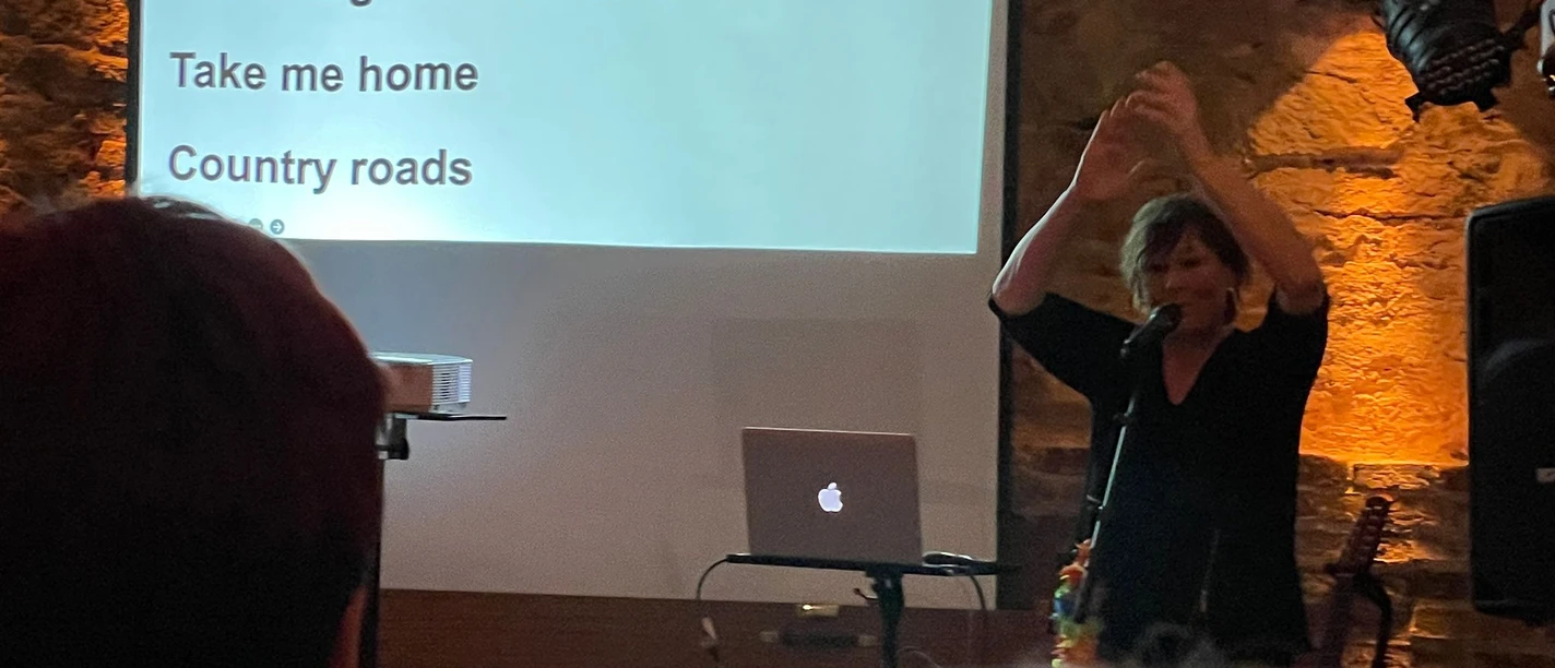 IMG-20240410-WA0008.jpg Frau mit Mikrofon vor Leinwand mit Text 'Da sprach der alte Häuptling der Indianer Wild ist der Westen schwer ist der Beruf 2x' vor Publikum in dunklem Raum