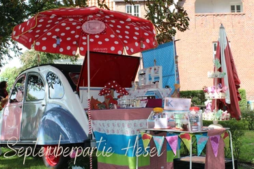 Septemberpartie Hesel.jpg Bunter Marktstand mit rotem Sonnenschirm, Oldtimer und handgefertigten Waren im Park von Hesel.
