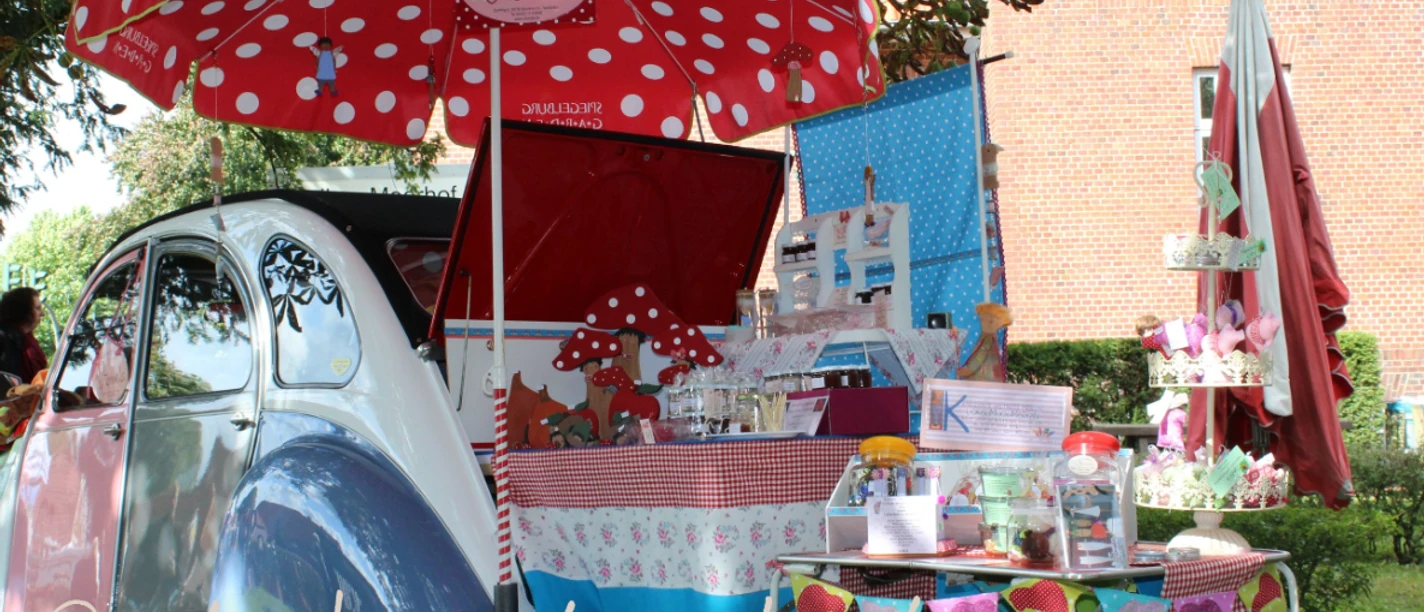 Septemberpartie Hesel.jpg Bunter Marktstand mit rotem Sonnenschirm, Oldtimer und handgefertigten Waren im Park von Hesel.