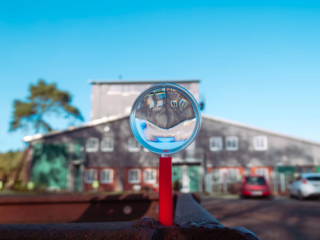 Ostersuche MoorIZ Ahlenmoor Lupe mit rotem Griff fokussiert den Blick auf das MoorInformationsZentrum, das sich unter klarem, blauem Himmel spiegelt.