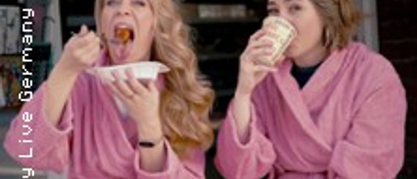 Zwei Frauen in rosa Bademänteln sitzen draußen, essen und trinken unter dem Schriftzug 1A B-WARE