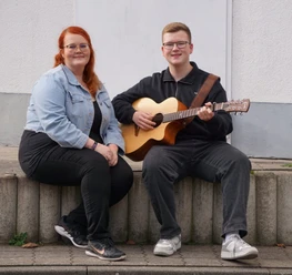 Duo Klangvoll.jpeg Zwei Personen sitzen auf einer niedrigen Mauer, eine spielt eine akustische Gitarre, die andere sitzt daneben mit verschränkten Händen.