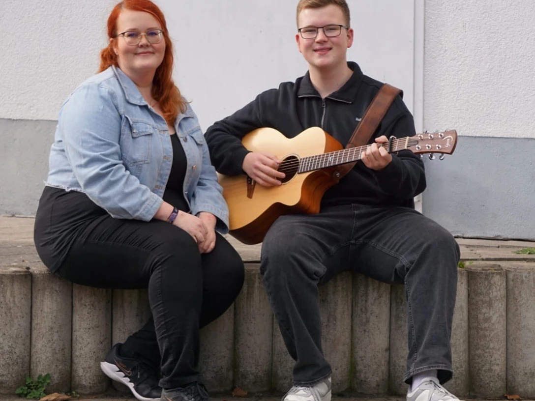 Duo Klangvoll.jpeg Zwei Personen sitzen auf einer niedrigen Mauer, eine spielt eine akustische Gitarre, die andere sitzt daneben mit verschränkten Händen.