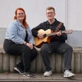 Duo Klangvoll.jpeg Zwei Personen sitzen auf einer niedrigen Mauer, eine spielt eine akustische Gitarre, die andere sitzt daneben mit verschränkten Händen.