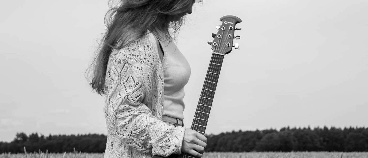Künstlerin.jpg Frau mit langem Haar und Strickjacke steht in einem Getreidefeld und hält eine Akustikgitarre im Stehen