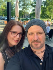 Musiker.jpeg Zwei Personen sitzen draußen an einem Tisch in einem belebten Biergarten mit Bäumen und geparkten Autos im Hintergrund