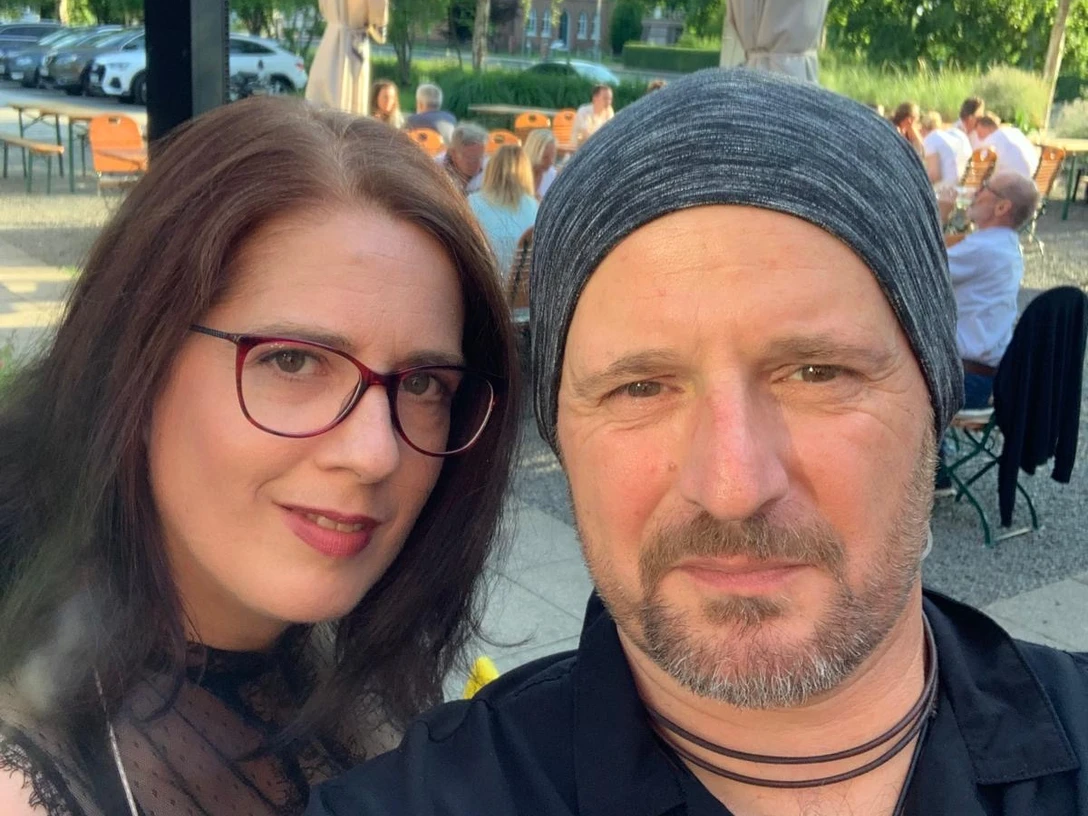 Musiker.jpeg Zwei Personen sitzen draußen an einem Tisch in einem belebten Biergarten mit Bäumen und geparkten Autos im Hintergrund