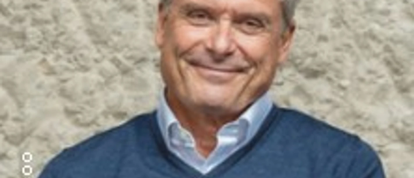 Älterer Mann mit grauem Haar sitzt lächelnd vor heller Steinwand in blauem Pullover.