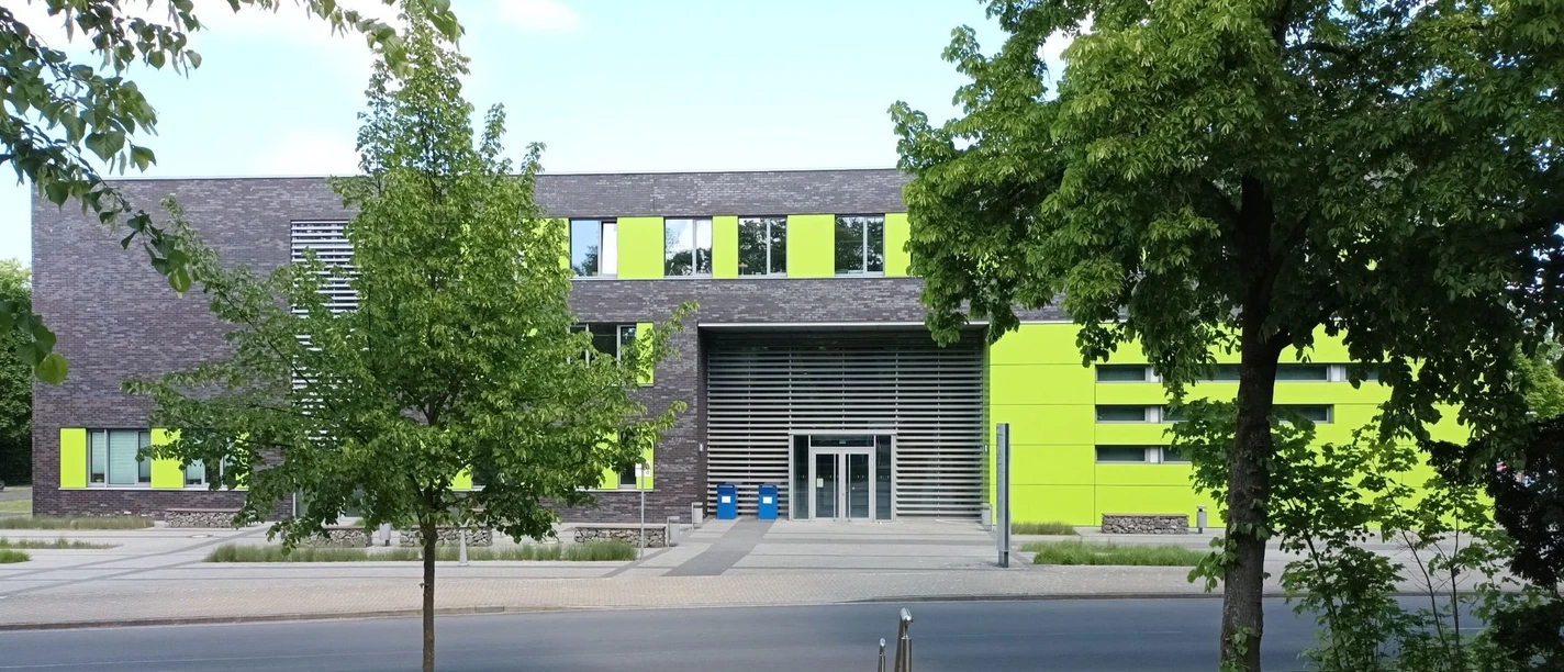 Ostfalia Hochschule_C-Gebaeude_HD_16zu9.jpg Modernes Hochschulgebäude mit grauer Fassade und leuchtend grünen Akzenten zwischen Bäumen.