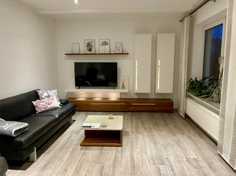 Ferienwohnung "Linde 28" Helles Wohnzimmer mit Sofa, Couchtisch, TV an Wandmöbel, Pflanzen am Fenster