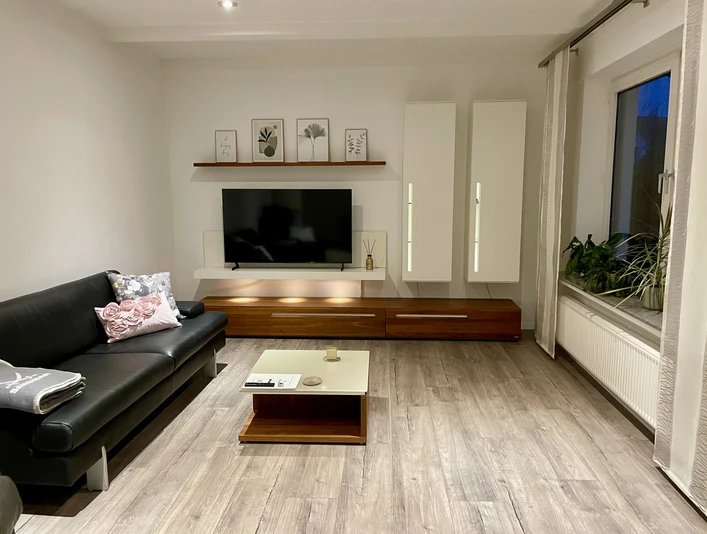 Ferienwohnung "Linde 28" Helles Wohnzimmer mit Sofa, Couchtisch, TV an Wandmöbel, Pflanzen am Fenster