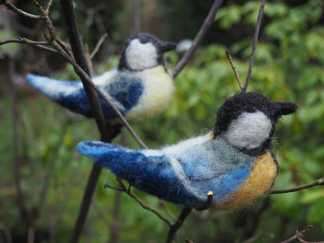 gefilzte Meisen Zwei handgefilzte Meisen in leuchtenden Farben auf Zweigen, umgeben von frischem Grün.