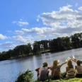 lopausee-amelinghausen-freizeit-erholung-sommer.jpg.png Eine Gruppe junger Menschen sitzt am grünen Ufer des Lopausees in Amelinghausen und genießt den Blick auf das Wasser unter blauem Sommerhimmel.