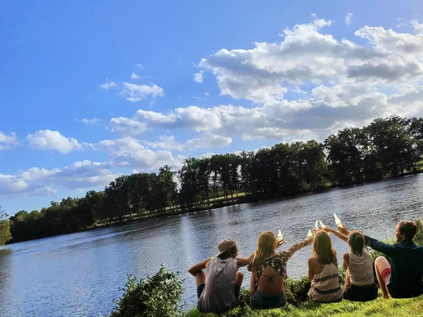 lopausee-amelinghausen-freizeit-erholung-sommer.jpg.png Eine Gruppe junger Menschen sitzt am grünen Ufer des Lopausees in Amelinghausen und genießt den Blick auf das Wasser unter blauem Sommerhimmel.