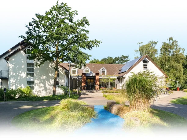 Modernes Hotelgebäude mit hellem Putz, Eingangsbereich, Grünflächen und kleinem Wasserlauf