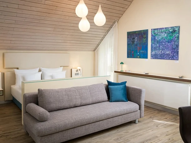 Modernes Hotelzimmer mit Doppelbett, Sofa, Holzdecke, Kunst an der Wand und Tageslicht.