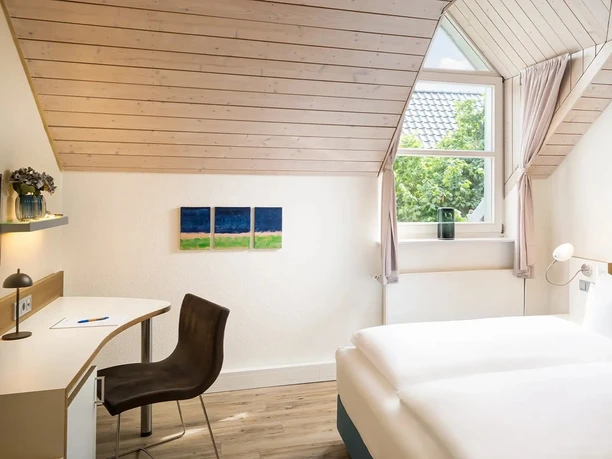 Helles Hotelzimmer mit Doppelbett, Schreibtisch, Holzdecke und Fenster unter Dachschräge.