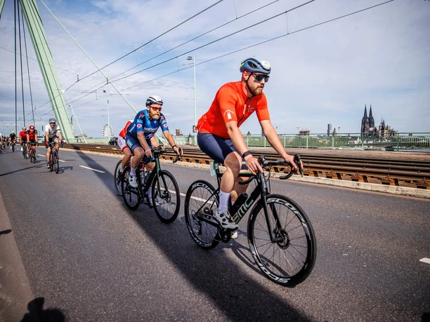 Rund um Köln Radfahrer auf einer Brücke mit Stadtsilhouette und Dom im Hintergrund