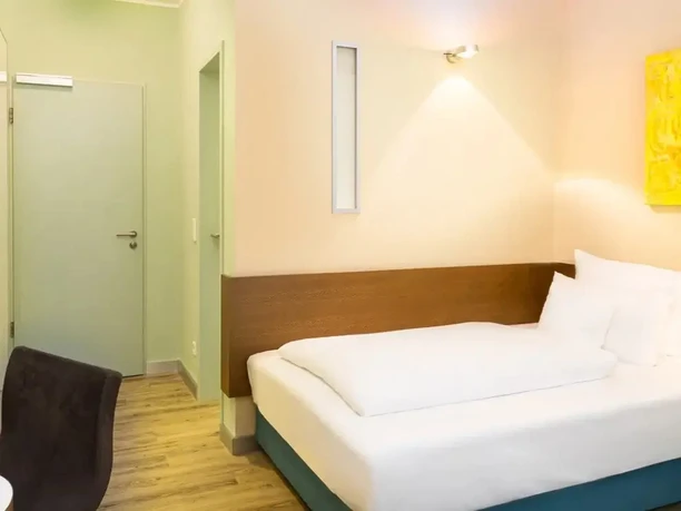 Modernes Hotelzimmer mit Doppelbett, hellen Wänden, Holzboden, Tür und Sessel.
