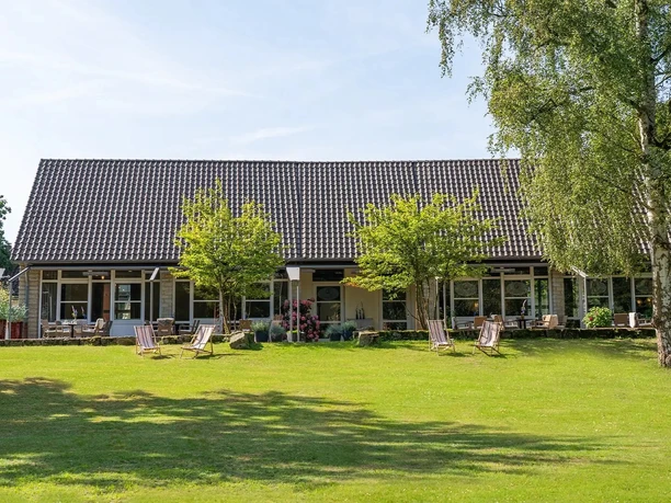 Flaches Hotelgebäude mit Terrasse, Liegestühlen und großem Garten mit Bäumen