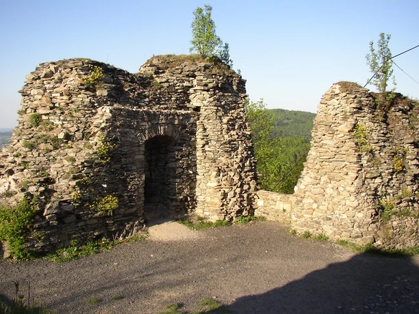 Überreste der Bastion in Tolštejn Ruinen alter Steinmauern mit Bäumen auf der Spitze, vor bewaldeten Hügeln.