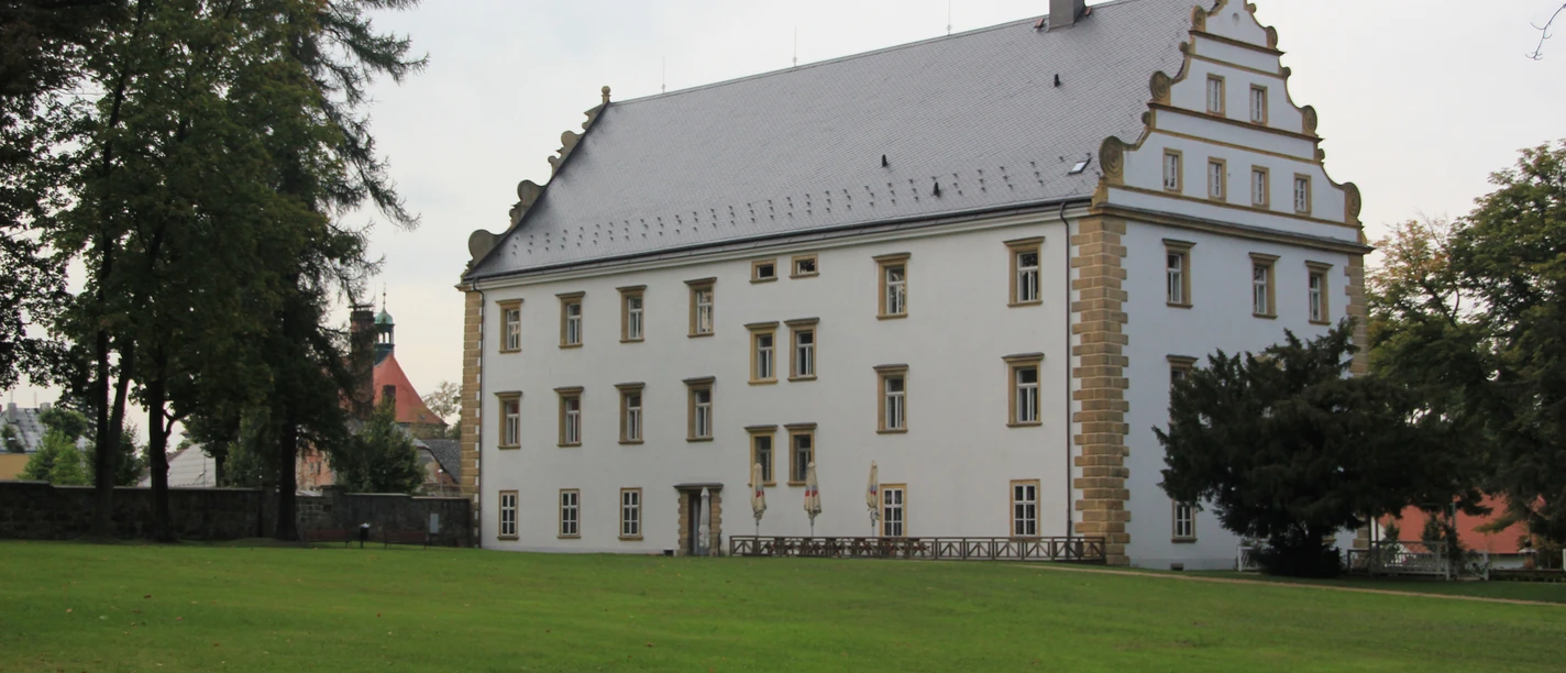 Weißes Schloss mit Ziergiebel, umgeben von einer Rasenfläche und Bäumen.