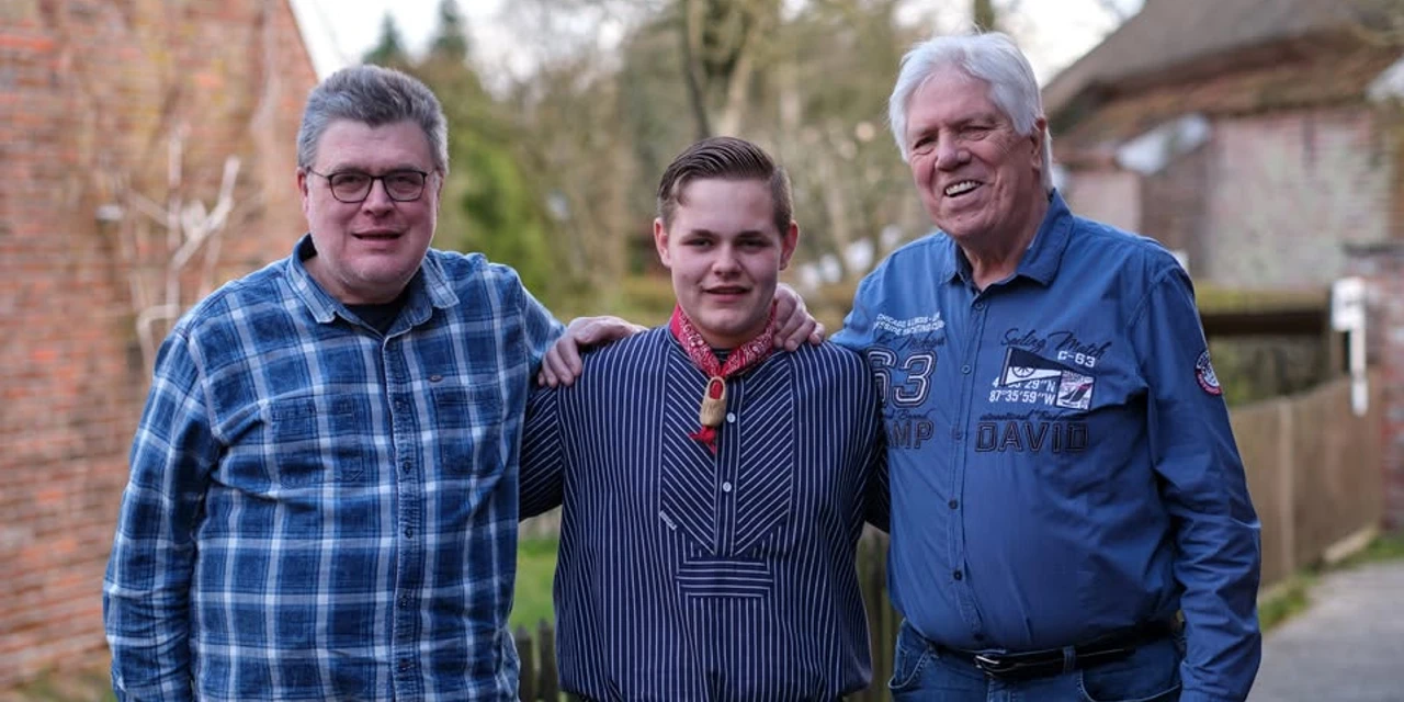 Hannes Flesner Trio.jpg Drei Männer stehen nebeneinander vor einem ländlichen Hintergrund, der mittlere trägt ein gestreiftes Hemd mit Halstuch, die beiden äußeren Männer tragen langärmlige Hemden, einer kariert, der andere mit Aufdrucken.