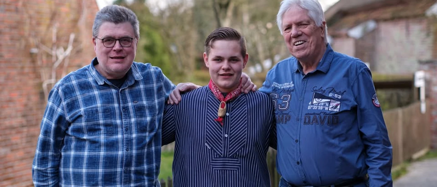 Hannes Flesner Trio.jpg Drei Männer stehen nebeneinander vor einem ländlichen Hintergrund, der mittlere trägt ein gestreiftes Hemd mit Halstuch, die beiden äußeren Männer tragen langärmlige Hemden, einer kariert, der andere mit Aufdrucken.