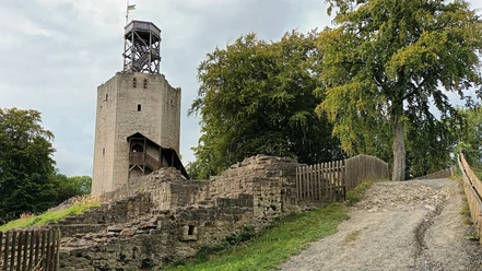 Burgfried Lichtenberg (33)_HD_16zu9.jpg Steinerner Burgturm mit hölzernem Aufsatz, umgeben von Mauerresten und Bäumen unter grauem Himmel.