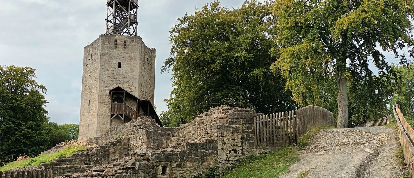 Burgfried Lichtenberg (33)_HD_16zu9.jpg Steinerner Burgturm mit hölzernem Aufsatz, umgeben von Mauerresten und Bäumen unter grauem Himmel.