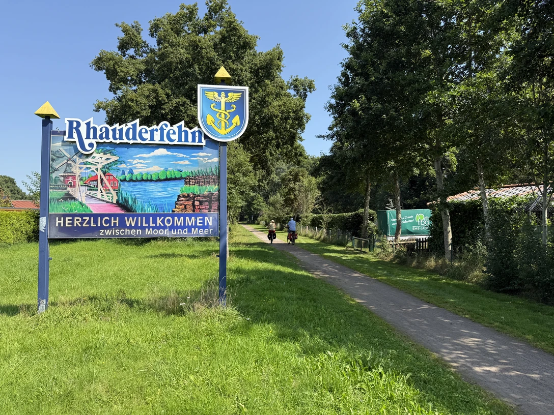 IMG_9712.JPG Willkommensschild mit dem Schriftzug 'Rhauderfehn' und 'Herzlich willkommen zwischen Moor und Meer' neben einem Weg mit zwei Radfahrern und Bäumen im Hintergrund