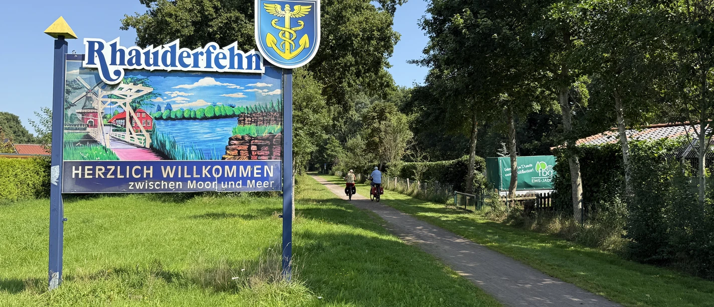 IMG_9712.JPG Willkommensschild mit dem Schriftzug 'Rhauderfehn' und 'Herzlich willkommen zwischen Moor und Meer' neben einem Weg mit zwei Radfahrern und Bäumen im Hintergrund