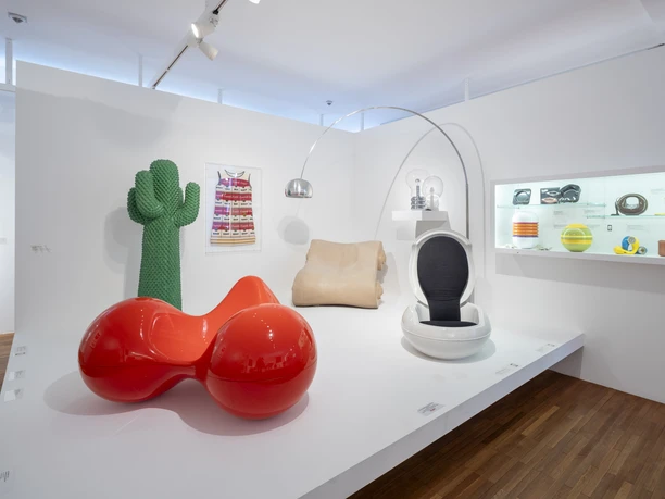 Museum für Angewandte Kunst Köln Ausstellungsraum mit farbigen Designobjekten wie rotem Sitzmöbel, Kaktusfigur und Retro-Stuhl.