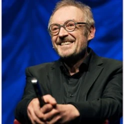 Älterer Mann mit Brille lächelnd auf Bühne und haltend Mikrofon vor blauem Hintergrund