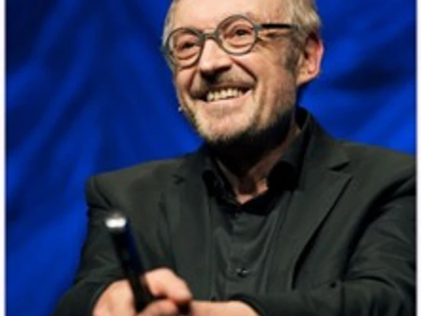 Älterer Mann mit Brille lächelnd auf Bühne und haltend Mikrofon vor blauem Hintergrund