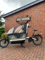 Auguste.jpg Lastenrad mit Wetterschutzplane vor roter Backsteinwand und Harley-Davidson-Logo.