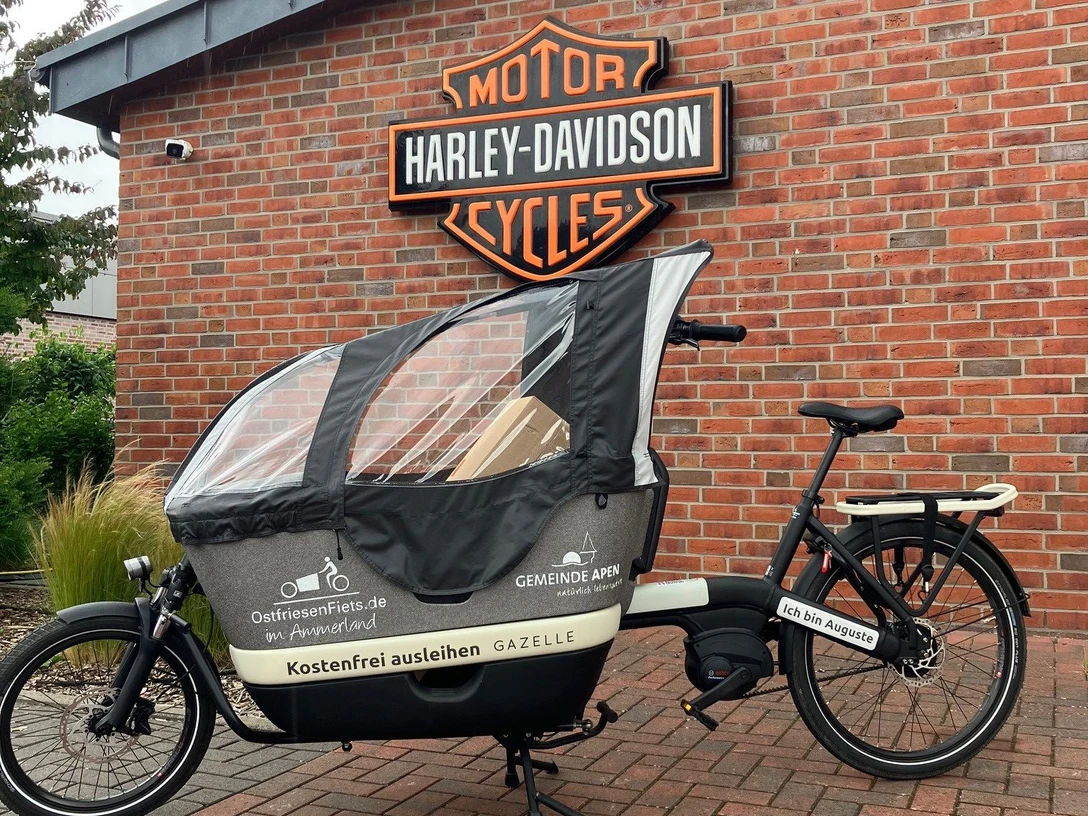 Auguste.jpg Lastenrad mit Wetterschutzplane vor roter Backsteinwand und Harley-Davidson-Logo.