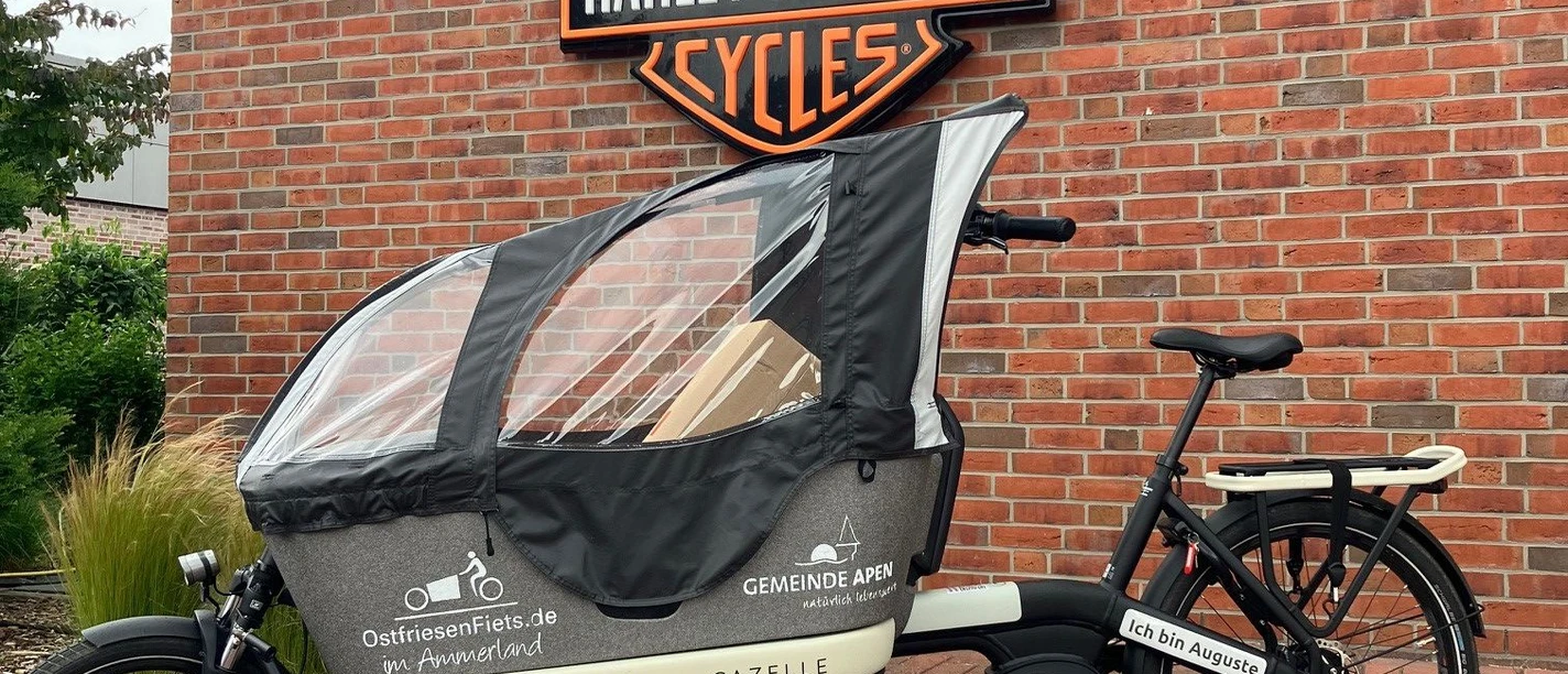 Auguste.jpg Lastenrad mit Wetterschutzplane vor roter Backsteinwand und Harley-Davidson-Logo.