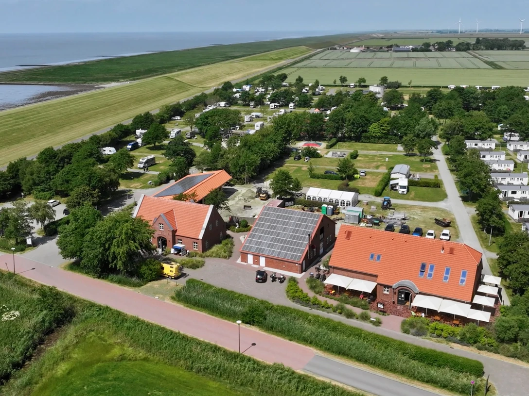 Camping am Deich Luftaufnahme eines Küstencampingplatzes hinter dem Deich mit Wohnwagen, Bäumen und Gebäuden.