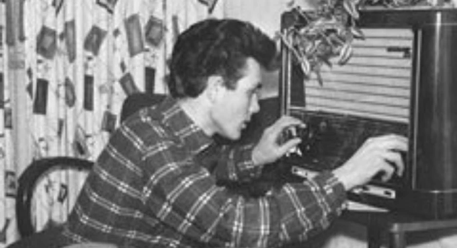 Das Lied vom Wirtschaftswunder Schwarzweißfoto eines jungen Mannes, der hockend an einem Röhrenradio die Einstellungen dreht.