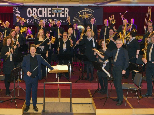Das Bild zeigt ein symphonisches Orchester auf der Bühne in Bad Zwischenahn. Die Musiker tragen formelle schwarze Kleidung und sind mit ihren Instrumenten bereit zum Auftakt eines Konzerts. Im Hintergrund ist ein Banner mit der Aufschrift "Orchester Bad Zwischenahn" zu sehen.