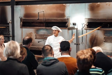 Öffentliches Abendbrot Bäckerei Ripken.jpg Ein Bäcker erklärt einer Gruppe vor großen Backöfen das traditionelle Brotbacken.