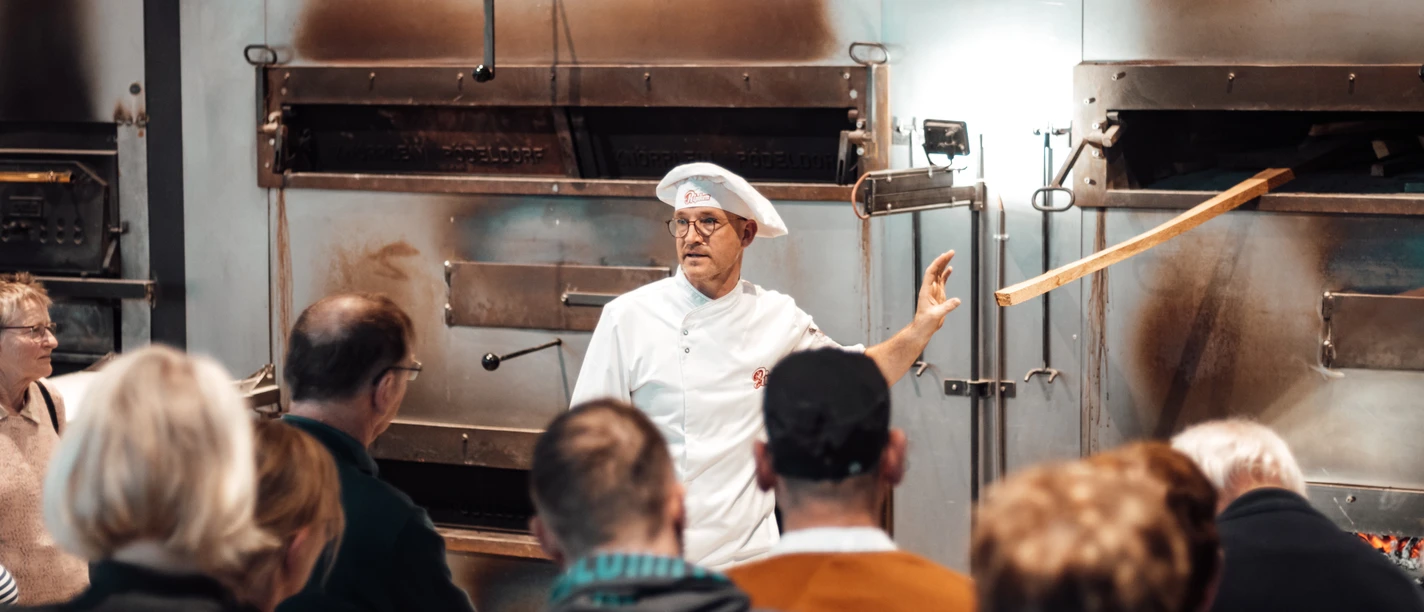 Öffentliches Abendbrot Bäckerei Ripken.jpg Ein Bäcker erklärt einer Gruppe vor großen Backöfen das traditionelle Brotbacken.
