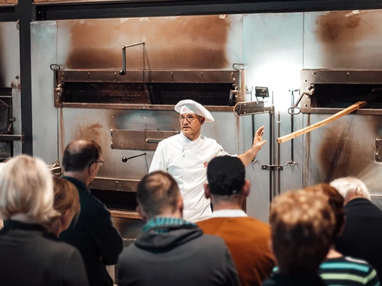 Öffentliches Abendbrot Bäckerei Ripken.jpg Ein Bäcker erklärt einer Gruppe vor großen Backöfen das traditionelle Brotbacken.
