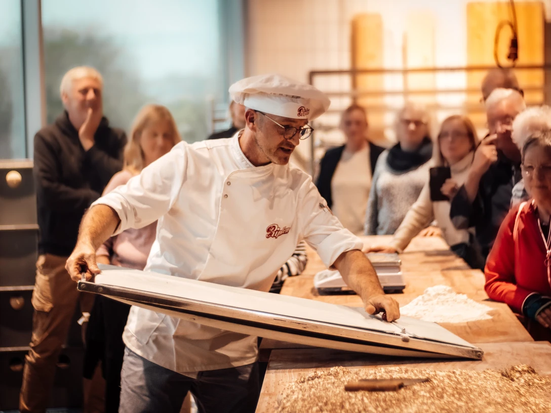 Öffentliches Abendbrot Bäckerei Ripken.jpg Bäcker in weißer Kleidung präsentiert Backvorgang vor interessierten Besucherinnen und Besuchern.