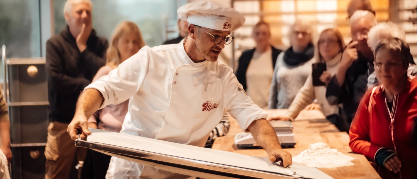 Öffentliches Abendbrot Bäckerei Ripken.jpg Bäcker in weißer Kleidung präsentiert Backvorgang vor interessierten Besucherinnen und Besuchern.