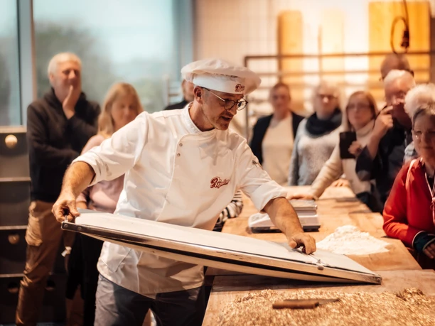 Öffentliches Abendbrot Bäckerei Ripken.jpg Bäcker in weißer Kleidung präsentiert Backvorgang vor interessierten Besucherinnen und Besuchern.