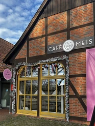 Außenansicht (Winter) (2) Fachwerkfassade des Café Mels mit winterlicher Dekoration und gelben Fenstern unter blauem Himmel.